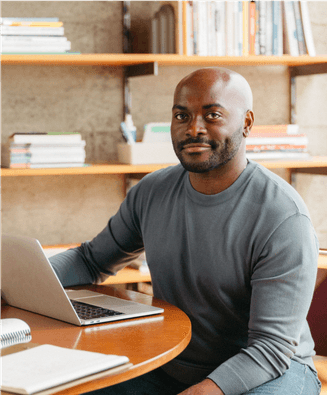 Man posing at a table with laptop and bookshelf